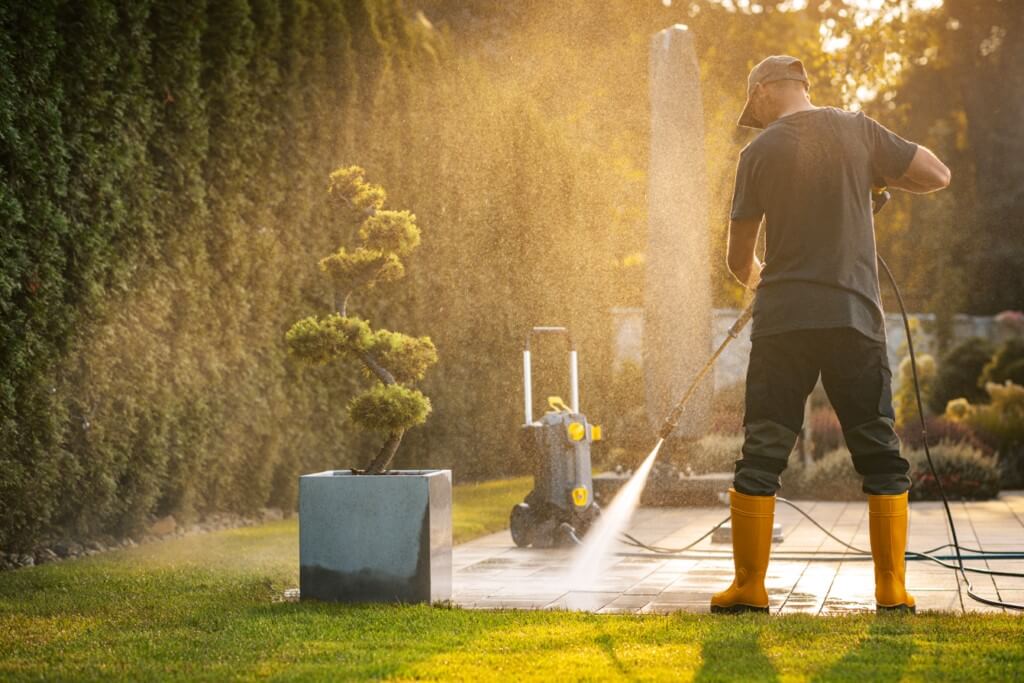 Homem de costas usando uma lavadora de alta pressão ou mangueira para limpar o piso de um quintal ensolarado. Ele veste botas amarelas e boné, direcionando o jato de água para as lajetas próximas a um vaso de planta decorativa e um gramado verde.