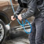 Close de um homem utilizando uma lavadora de alta pressão ou mangueira para remover a sujeira das rodas e da lateral de um carro marrom. Ele segura a ponteira da lavadora com as duas mãos, focando o jato de água diretamente no pneu dianteiro.